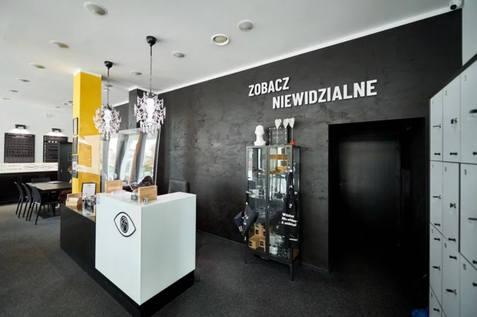 Recepcja Black Word. Wystrój przywodzi na myśl tryb zwiększonego kontrastu barwnego. Ściany i podłoga czarne, sufit i biurko białe, słup żółty. Na ścianie napis Zabacz niewidzialne. Po lewej stronie szafki na klucz, po prawej lada do obsługi gości. Na wprost czarny korytarz do dalszych pomieszczeń.