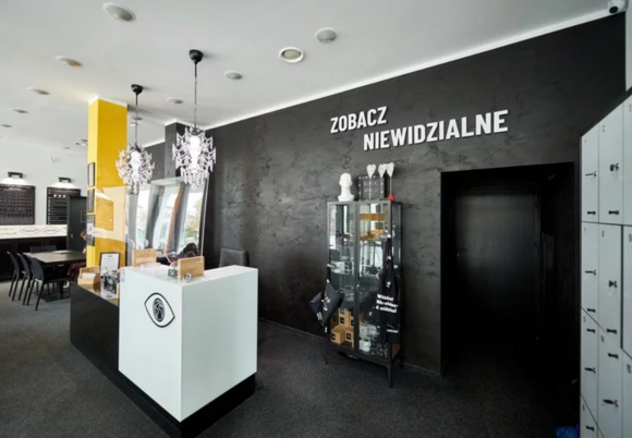 Recepcja Black Word. Wystrój przywodzi na myśl tryb zwiększonego kontrastu barwnego. Ściany i podłoga czarne, sufit i biurko białe, słup żółty. Na ścianie napis Zabacz niewidzialne. Po lewej stronie szafki na klucz, po prawej lada do obsługi gości. Na wprost czarny korytarz do dalszych pomieszczeń.